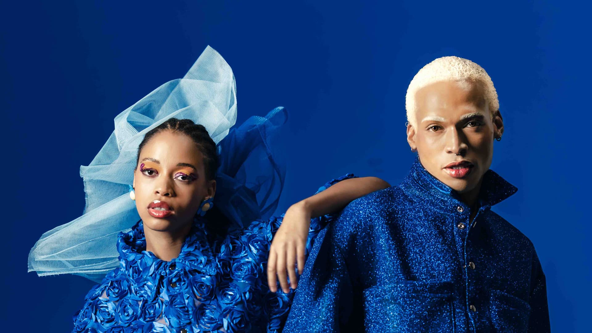 Two models in floral and glittery blue outfits pose against a solid blue background.
