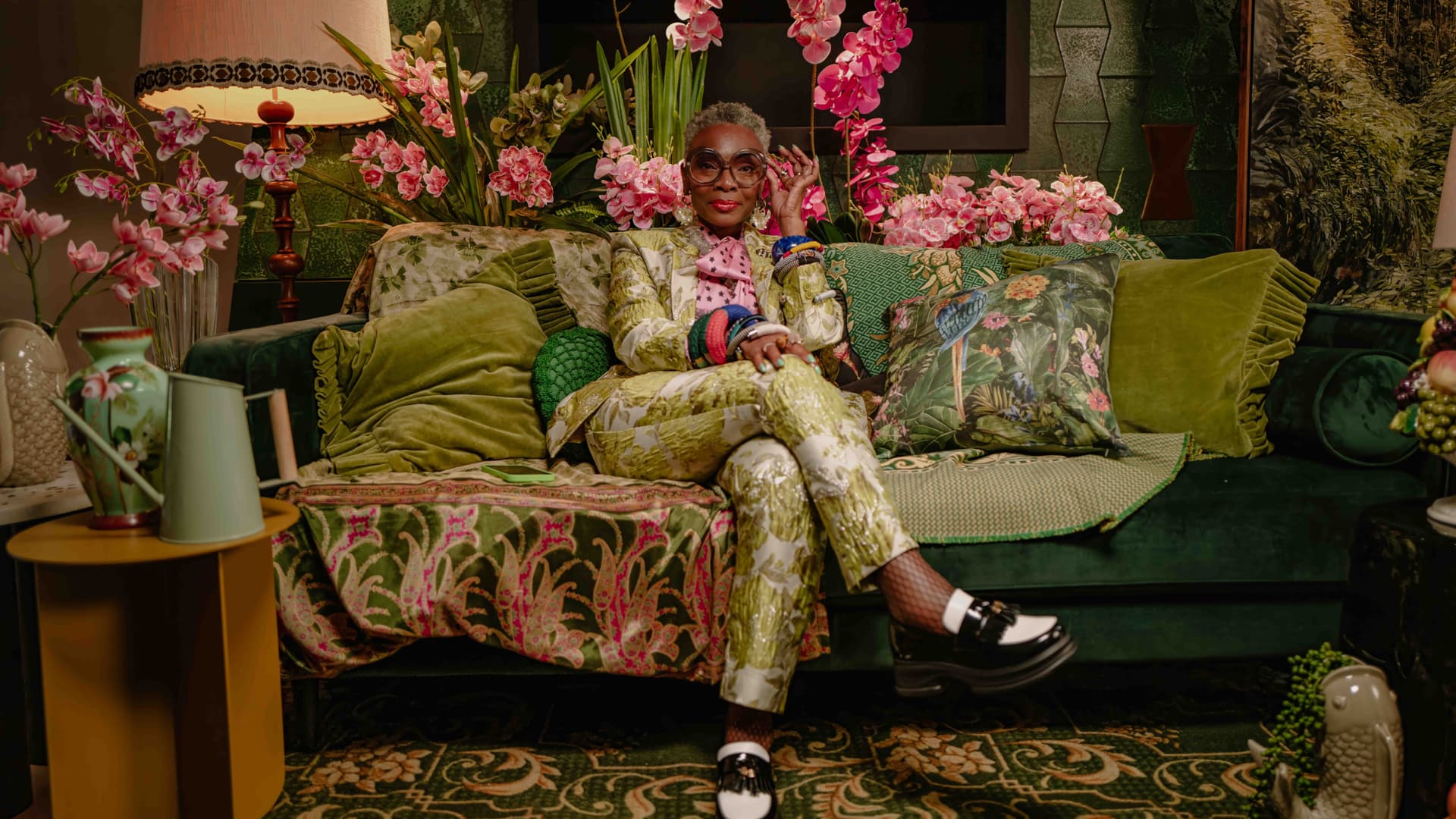 Stylish woman in green patterned suit sits on velvet sofa surrounded by vibrant pink orchids.