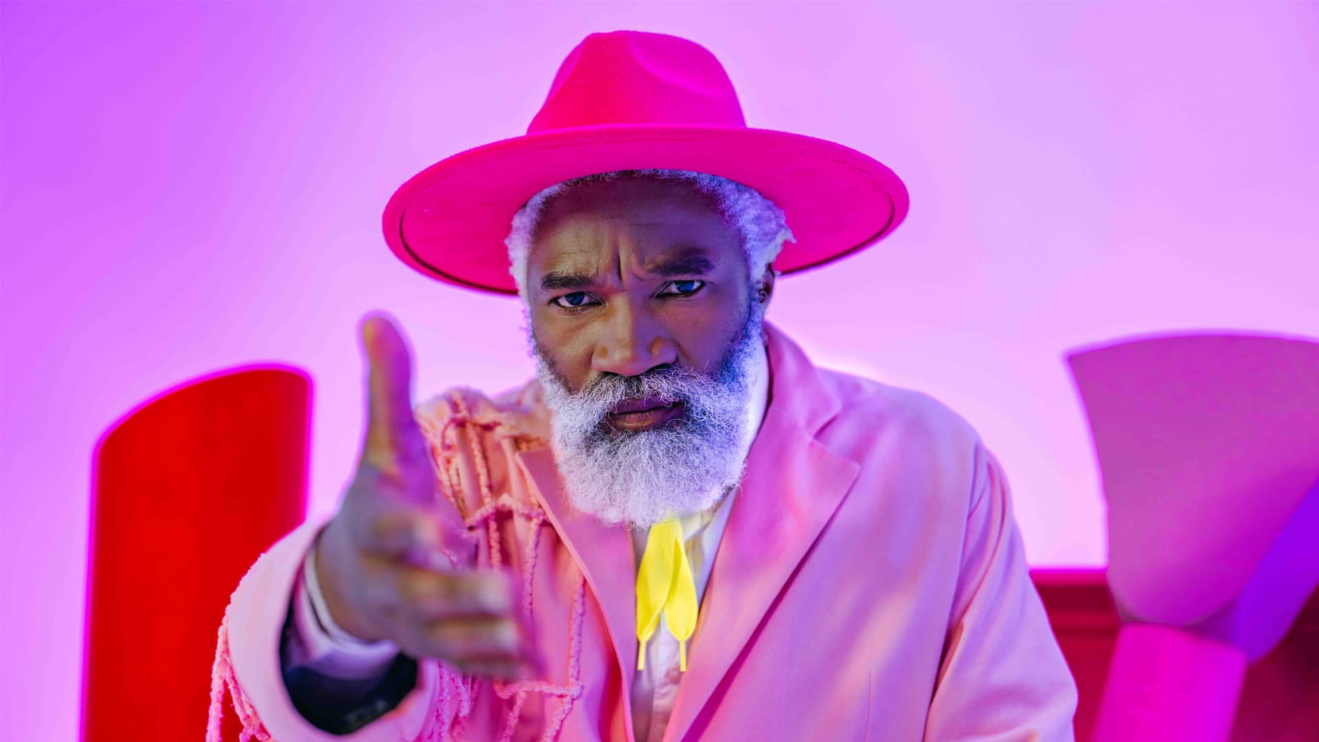 Stylish older Black man in a pink hat and suit reaching out against pink lighting.