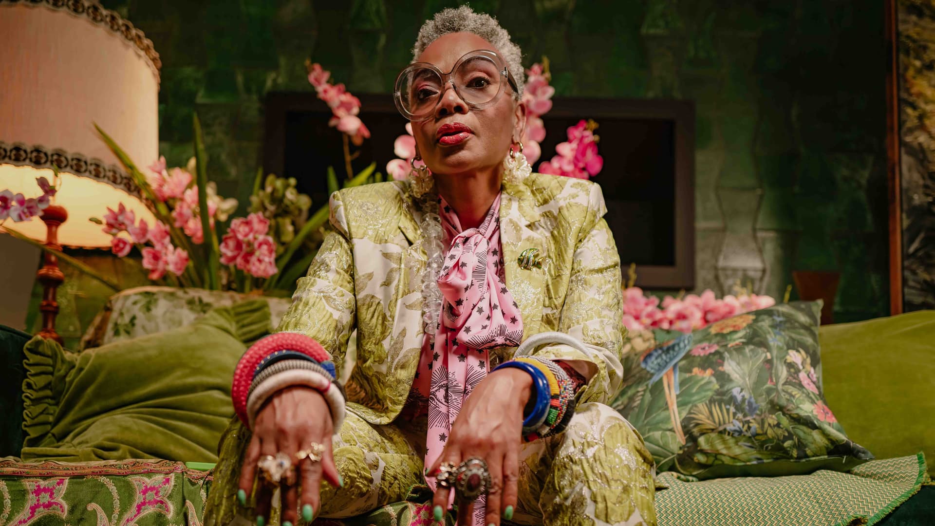 Stylish older Black woman in a green suit and large glasses on a green couch.