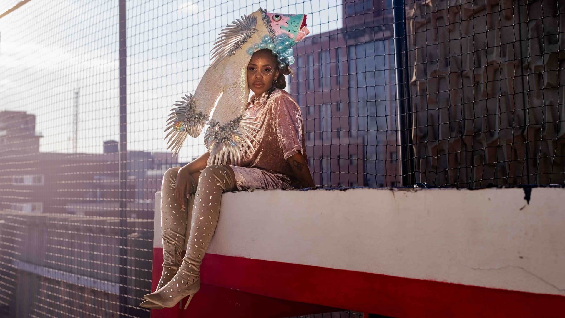Woman wearing an elaborate fish headpiece and sparkly boots sits on a city rooftop ledge.