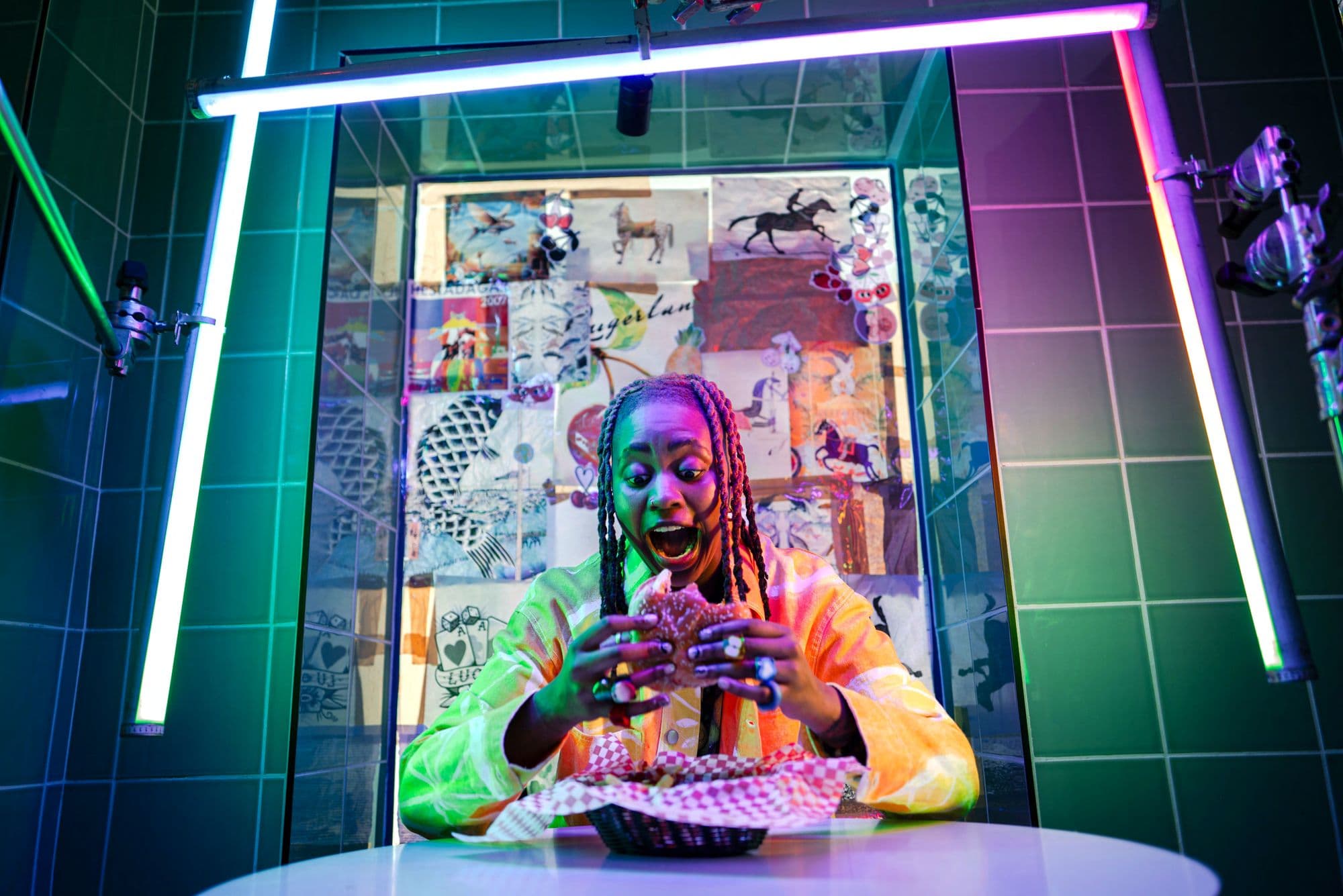 Excited person with braids about to eat a burger under vibrant, colorful neon lights.