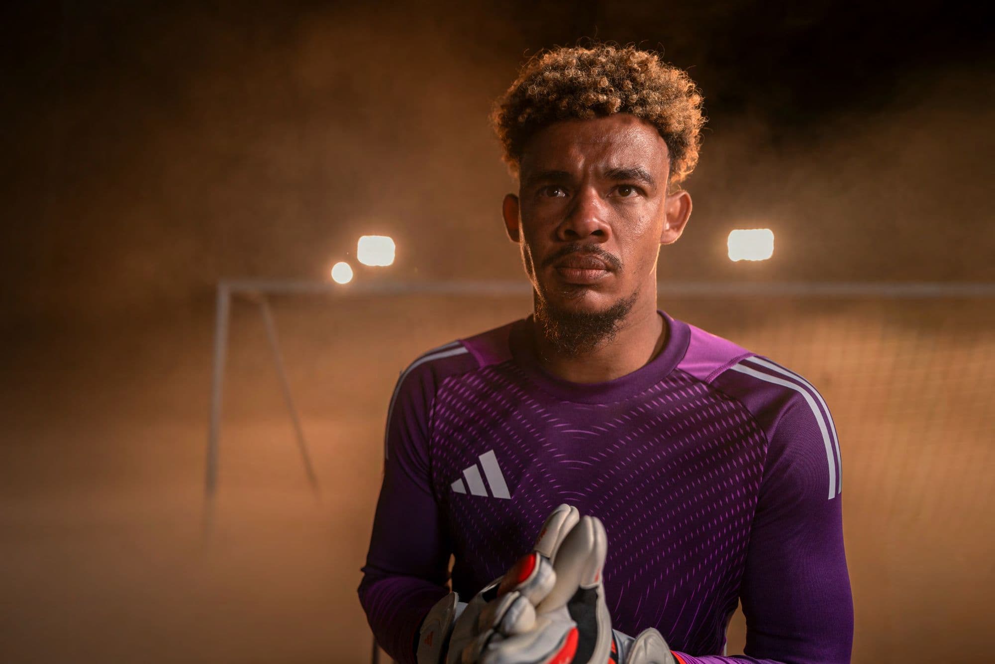 Focused soccer goalkeeper in a purple jersey stands before a goal in a smoky stadium.