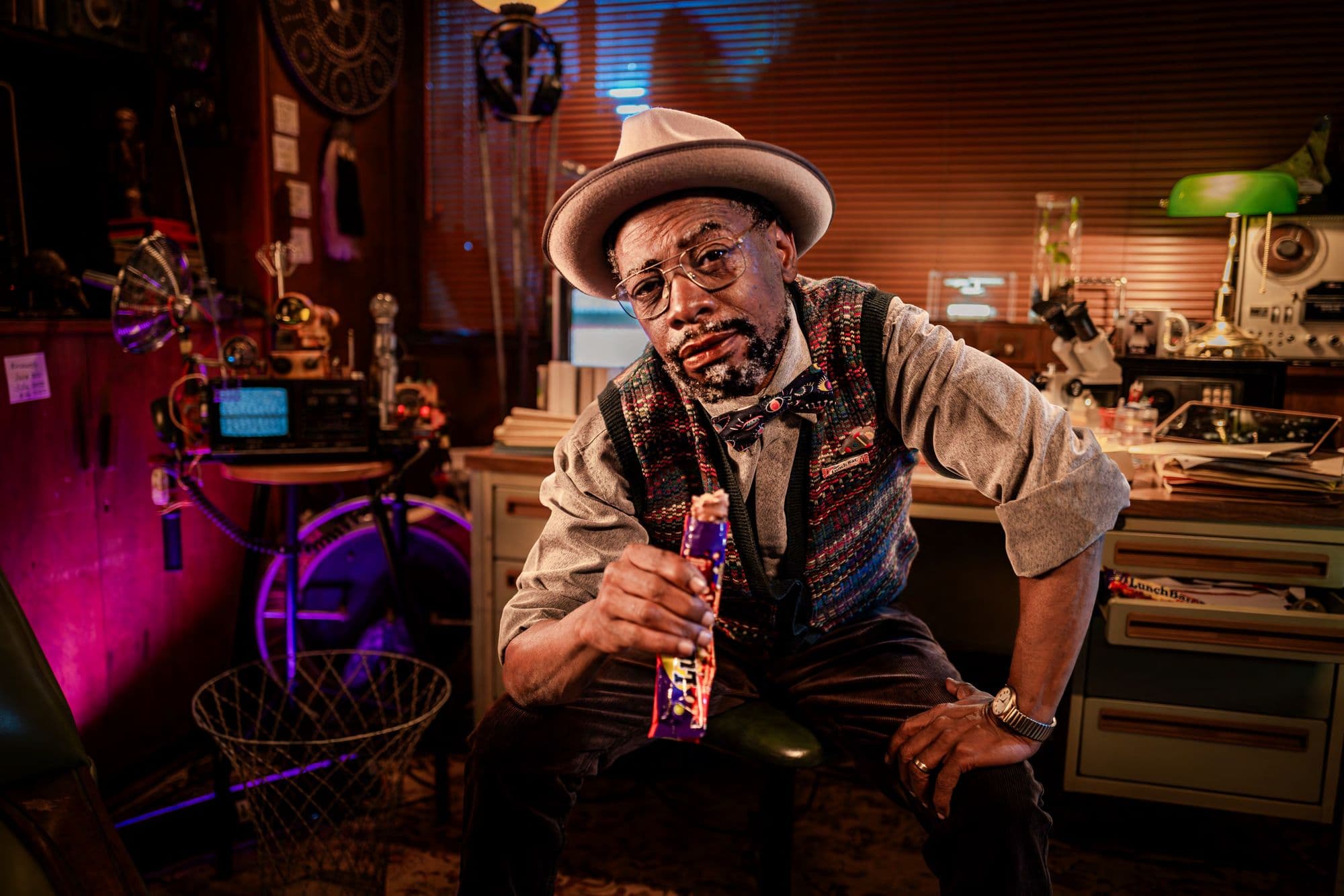 Older man in a fedora and vest holding a chocolate bar in a cluttered workshop.