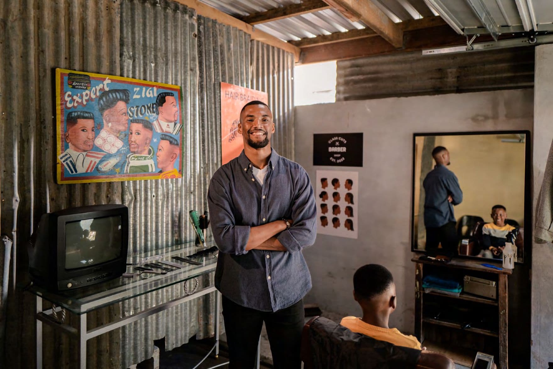 Smiling barber stands with arms crossed in a rustic barbershop with corrugated metal walls.