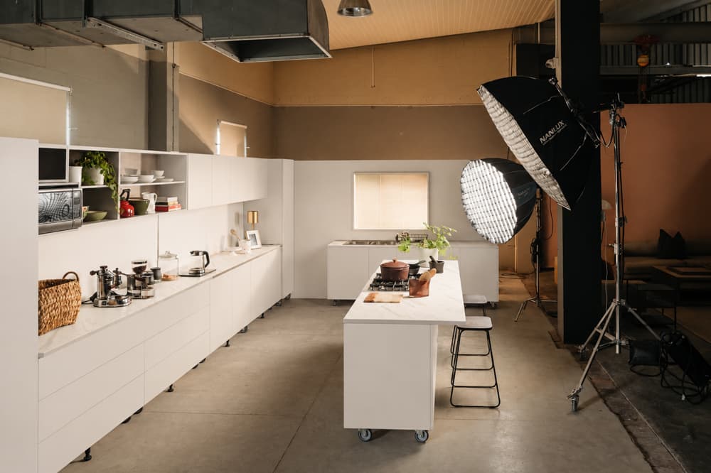 Modern white kitchen set in an industrial studio with large professional softbox lighting equipment.
