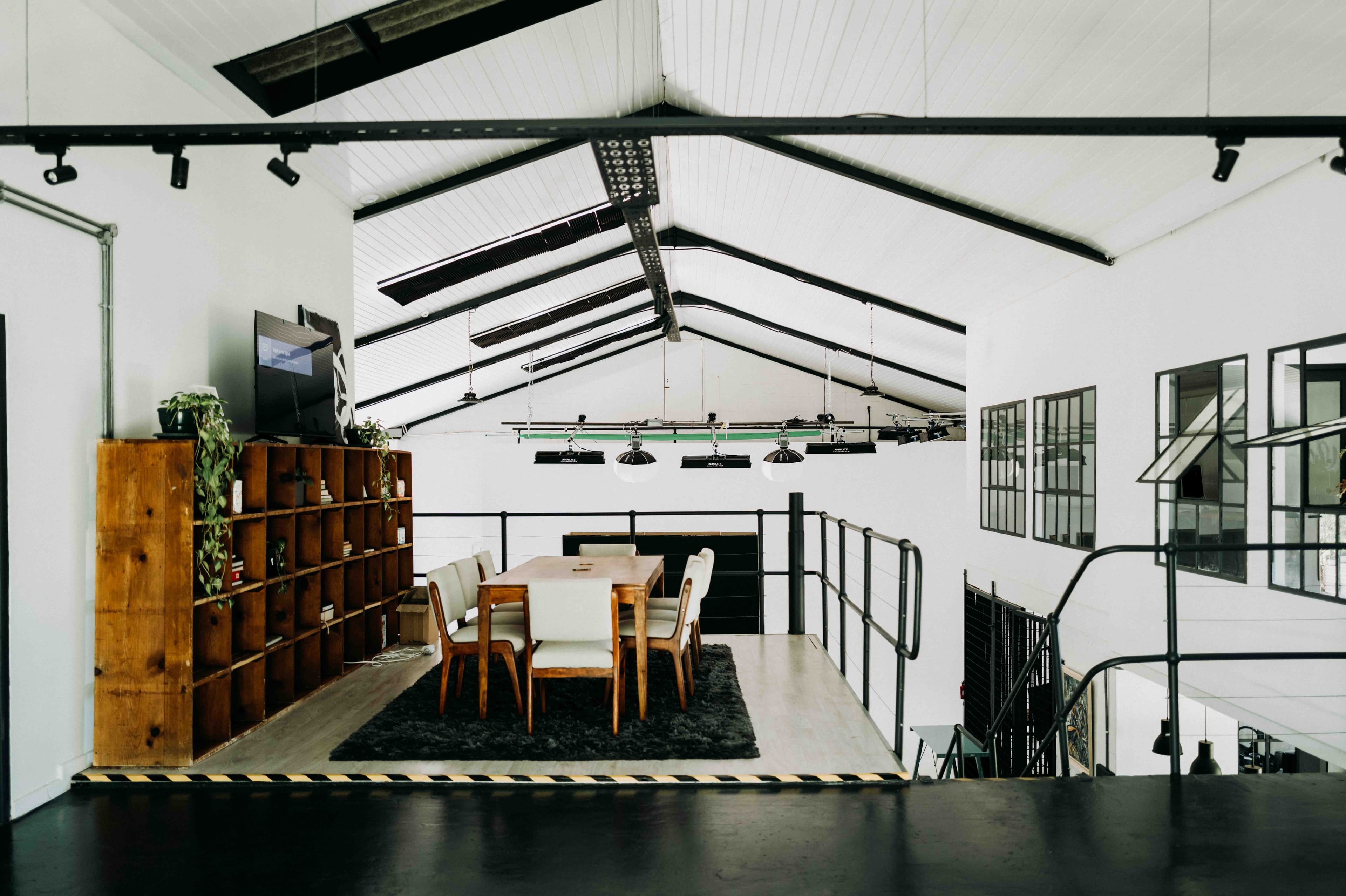 Modern mezzanine studio with a wooden table, large bookshelf, and high pitched industrial ceiling.