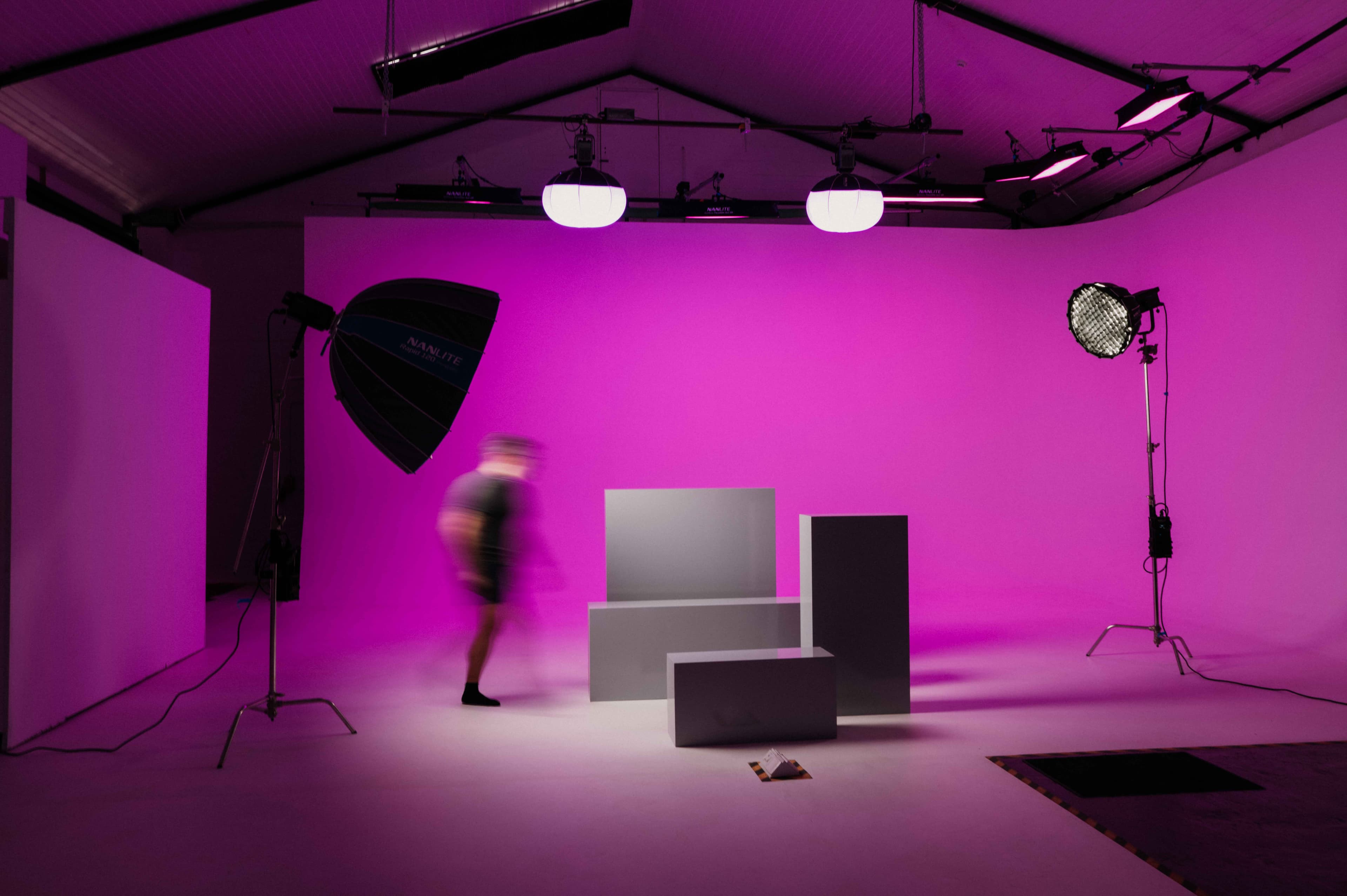 Professional studio featuring a pink backdrop, lighting equipment, grey pedestals, and a blurred person.